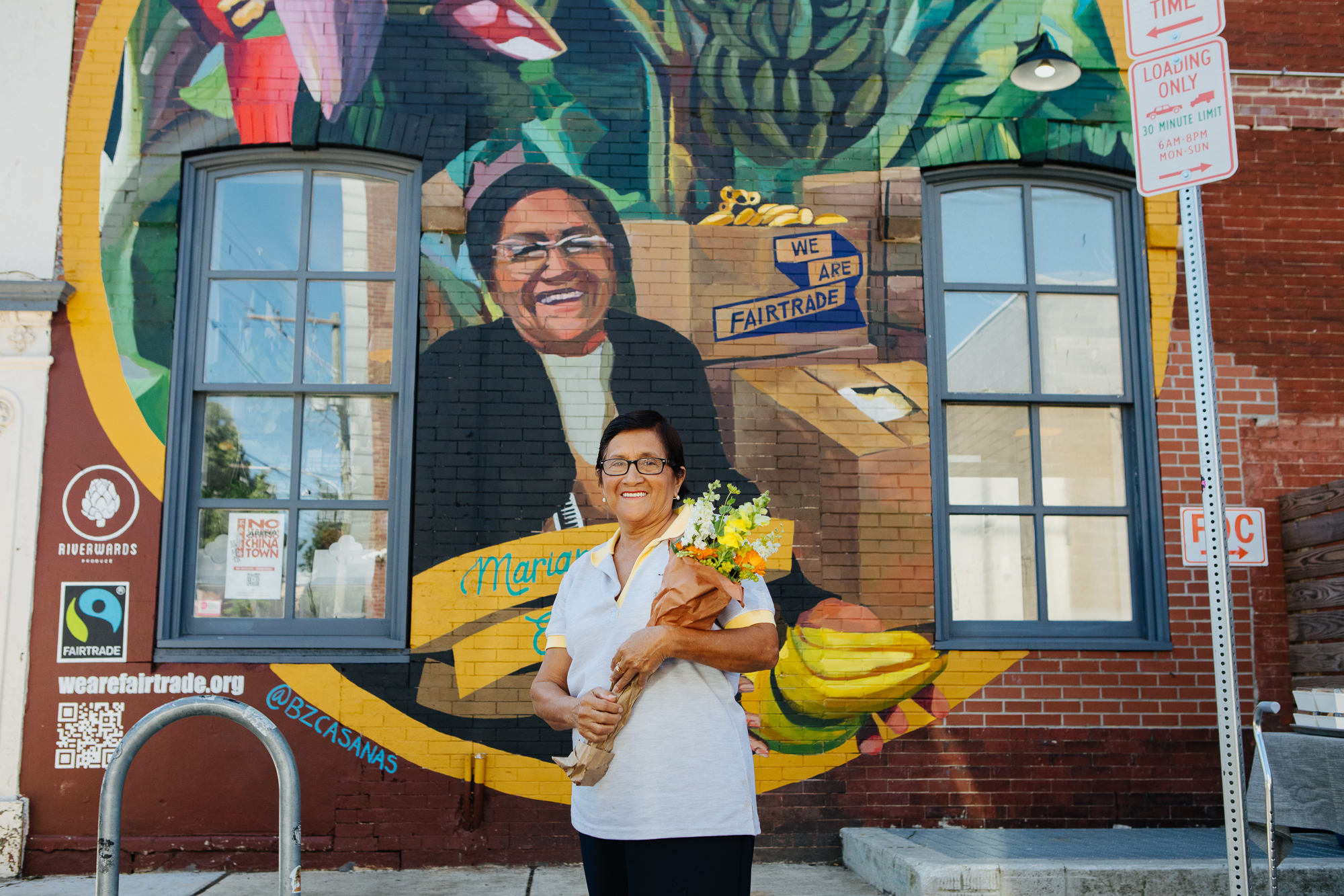 A woman with a bouquet of flowers stands in front of a mural on the side of a brick building that depicts the same woman